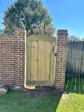 Pressure-treated wood arched gate with black iron hardware between red brick pillars beside a wood privacy fence