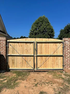 Double swing wood driveway gate with vertical boards, horizontal rails, diagonal bracing, and steel hinges between brick pillars