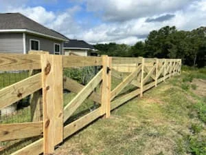 Three-rail pressure-treated wood ranch fence with square posts running along a grassy yard edge