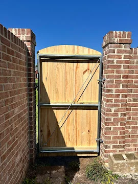 Arched-top wood gate with diagonal brace and black metal hinges set between red brick pillars with corbelled caps
