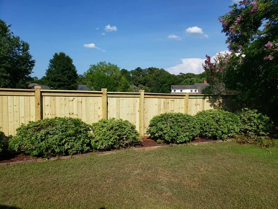A newly installed wood privacy fence running along the back yard of a Middle Georgia home, with red clay soil visible at the post bases and pine trees in the background.