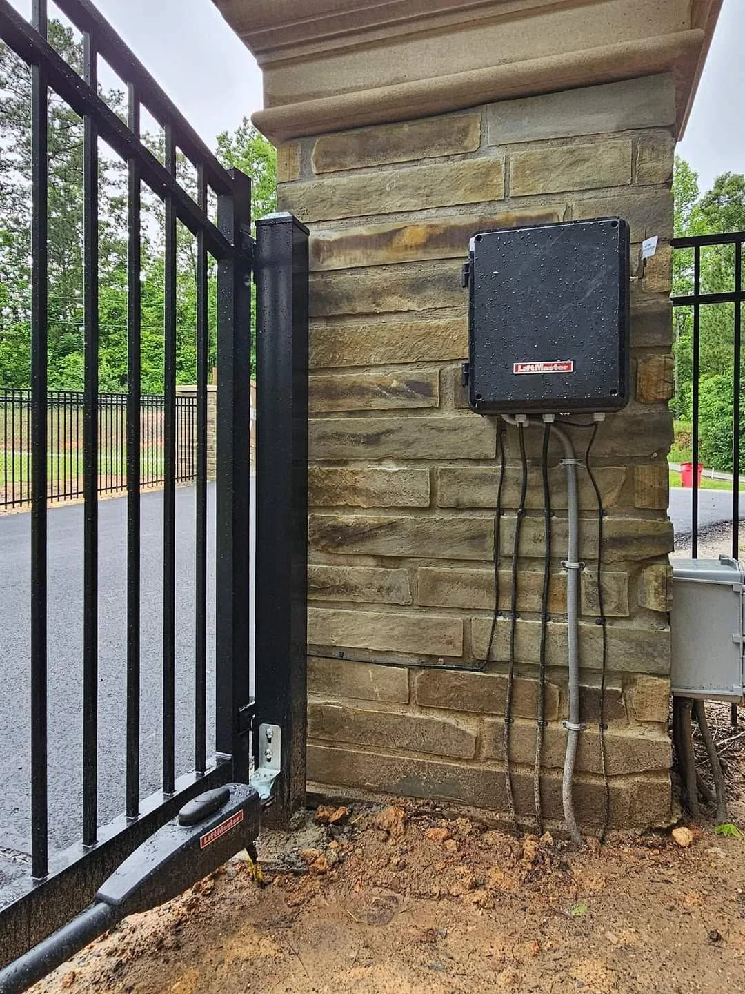 A residential driveway gate with an automatic opener installed on a post, photographed at a home in the Warner Robins, Georgia area. The operator unit, keypad pedestal, and swing gate are visible in the foreground with a residential fence line extending behind it.