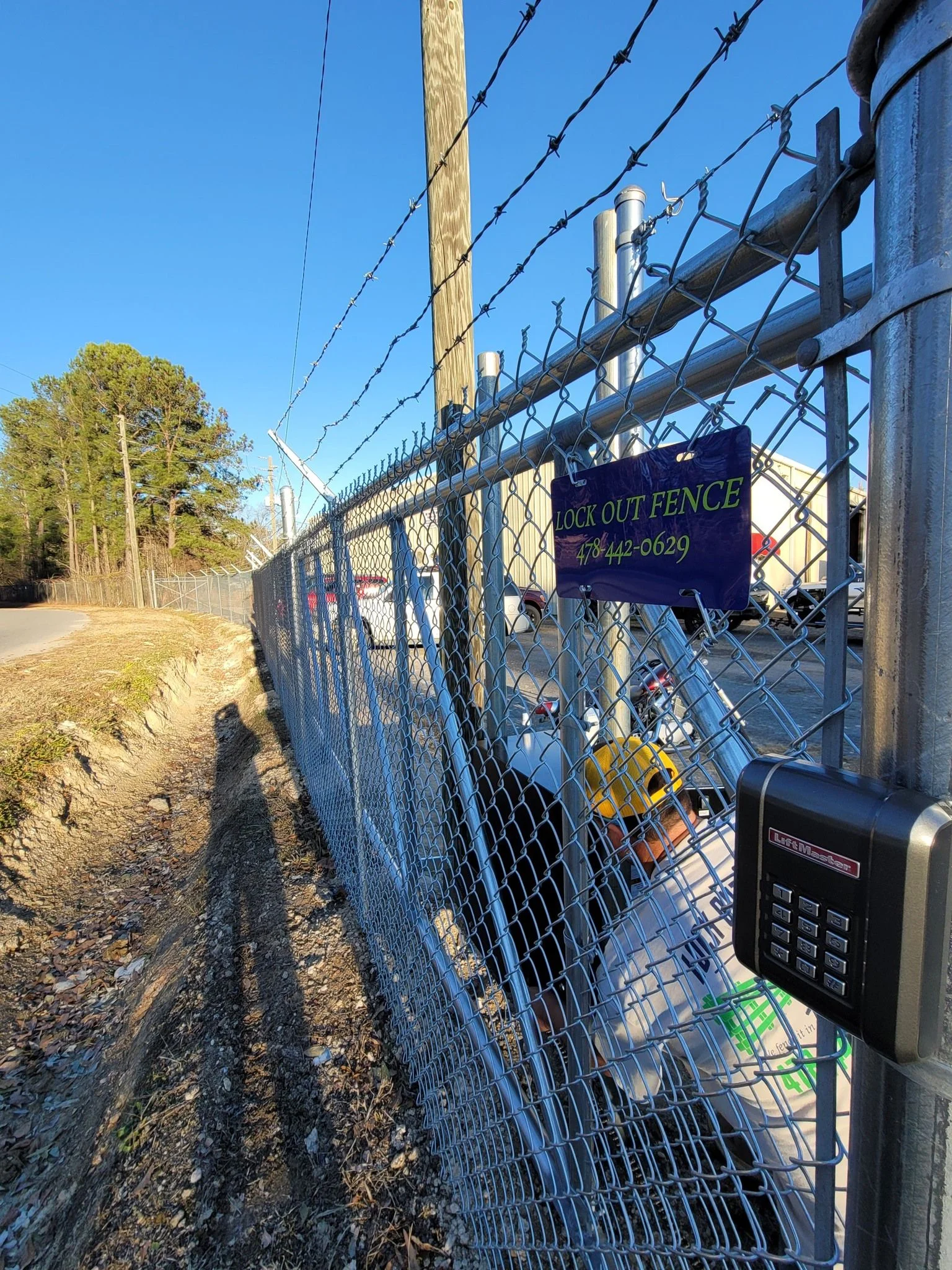 A residential driveway gate opener motor mounted on a post in Warner Robins, Georgia, with a technician from Lock Out Fence LLC inspecting the control panel and wiring. The gate is a swing-style fence gate set against a typical Middle Georgia residential property.